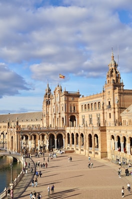 A grand architectural structure with ornate details and numerous arches extends into a large plaza. People are scattered throughout the open area, enjoying the sunny day. A Spanish flag waves atop one of the building's towers. The sky is partly cloudy, creating a pleasant atmosphere.