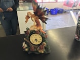 A decorative clock on a table features a fairy with long, flowing hair and large iridescent wings sitting atop it. The clock is adorned with roses and leaves. In the background, part of a person wearing overalls is visible, as well as some blue bins and red shopping carts.
