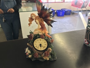 A decorative clock on a table features a fairy with long, flowing hair and large iridescent wings sitting atop it. The clock is adorned with roses and leaves. In the background, part of a person wearing overalls is visible, as well as some blue bins and red shopping carts.