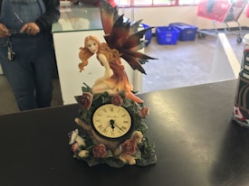 A decorative clock on a table features a fairy with long, flowing hair and large iridescent wings sitting atop it. The clock is adorned with roses and leaves. In the background, part of a person wearing overalls is visible, as well as some blue bins and red shopping carts.