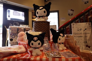 A display featuring multiple plush toys resembling a character with black cat-like attire, all adorned with a small pink skull on their foreheads. They are arranged on a checkered tablecloth, with one toy elevated on a wicker basket. Surrounding the toys are various items, including packaged products and a sign indicating the availability of a special room in the cafe. The background features posters, a window, and a staircase.