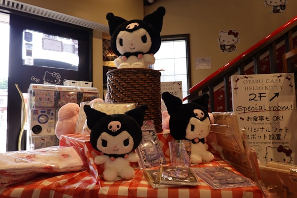 A display featuring multiple plush toys resembling a character with black cat-like attire, all adorned with a small pink skull on their foreheads. They are arranged on a checkered tablecloth, with one toy elevated on a wicker basket. Surrounding the toys are various items, including packaged products and a sign indicating the availability of a special room in the cafe. The background features posters, a window, and a staircase.