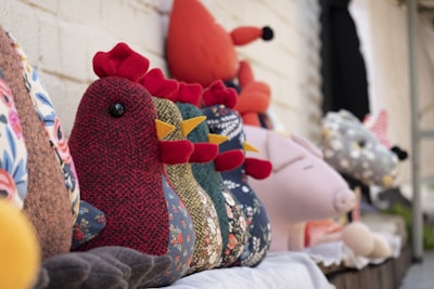 A collection of colorful, handmade stuffed animal toys is arranged in a line. The foreground features a rooster crafted from patterned fabric, displaying intricate textures and vibrant colors. Other animals, including a fox and a pig, are visible in the background, all made with similar attention to detail.