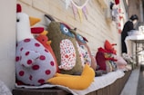 Several handmade plush toys, including a chicken with red polka-dotted fabric and an owl with colorful patterned fabric, are arranged on a wooden ledge. The background shows a light brick wall adorned with a string of patterned triangular flags. In the background, a person is standing near a table covered with a white cloth, appearing to be engaged in some activity.