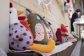 Several handmade plush toys, including a chicken with red polka-dotted fabric and an owl with colorful patterned fabric, are arranged on a wooden ledge. The background shows a light brick wall adorned with a string of patterned triangular flags. In the background, a person is standing near a table covered with a white cloth, appearing to be engaged in some activity.