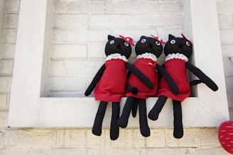 Three plush cat dolls wearing red dresses with white collars are sitting together on a white ledge. Each cat has button eyes, black fur, and a small red bow near one ear. The background consists of a light-colored brick wall.