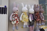 Several plush rabbit toys with long ears are hanging in a row against a wall. They are dressed in various colorful outfits: floral patterns, stripes, and solid colors. Each rabbit has soft, round features and is posed upright.