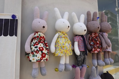 Several plush rabbit toys with long ears are hanging in a row against a wall. They are dressed in various colorful outfits: floral patterns, stripes, and solid colors. Each rabbit has soft, round features and is posed upright.