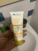 A hand is holding a tube of Garnier SkinActive face cleanser called Vitamine C Mousse Nettoyante Visage. The packaging is predominantly white and yellow with text in French. The background includes a bathroom setting with tiles and other hygiene products barely visible.