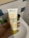 A hand is holding a tube of Garnier SkinActive face cleanser called Vitamine C Mousse Nettoyante Visage. The packaging is predominantly white and yellow with text in French. The background includes a bathroom setting with tiles and other hygiene products barely visible.