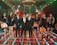 A group of eight people stands on an illuminated, colorful stage with overhead lighting. The men are dressed in black suits with orange ties, while the women wear black dresses and hold orange briefcases. The background includes digital screens displaying abstract designs, and the atmosphere is vibrant and celebratory.