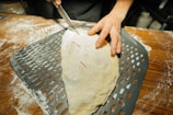 A person is using a knife to make slits in a dough-covered calzone or similar pastry placed on a perforated baking sheet. The surface is dusted with flour, and one of the hands shows visible flour marks.