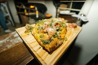 A rectangular wooden board holds a freshly baked pizza topped with green herbs, slices of thin meat, and melted cheese. The background is slightly blurred, suggesting a cozy kitchen or pizzeria environment.