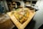 A rectangular wooden board holds a freshly baked pizza topped with green herbs, slices of thin meat, and melted cheese. The background is slightly blurred, suggesting a cozy kitchen or pizzeria environment.