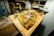 A rectangular wooden board holds a freshly baked pizza topped with green herbs, slices of thin meat, and melted cheese. The background is slightly blurred, suggesting a cozy kitchen or pizzeria environment.