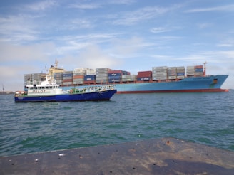 Two ships are seen on the water; a large cargo ship carrying stacked shipping containers and a smaller pilot boat alongside it. The sea appears calm under a partly cloudy sky.
