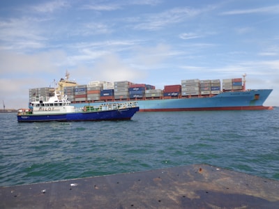 Two ships are seen on the water; a large cargo ship carrying stacked shipping containers and a smaller pilot boat alongside it. The sea appears calm under a partly cloudy sky.