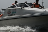 A motorboat labeled 'KAVERI 4' is cutting through the water at high speed, creating splashes at the front. Several people wearing bright orange life vests are on board, some visible through the boat's windows. The boat has insignia and flags on its side, hinting at an official or governmental purpose.