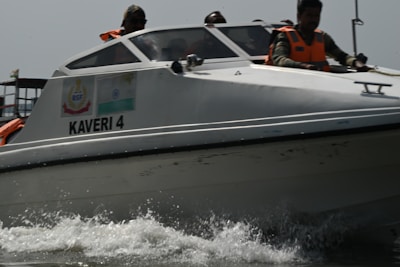A motorboat labeled 'KAVERI 4' is cutting through the water at high speed, creating splashes at the front. Several people wearing bright orange life vests are on board, some visible through the boat's windows. The boat has insignia and flags on its side, hinting at an official or governmental purpose.