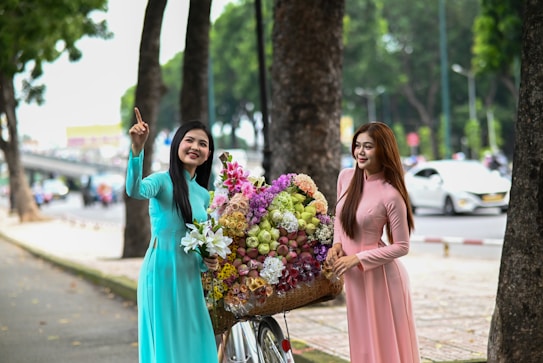 Two women in traditional dresses stand beside a bicycle laden with a large basket of colorful flowers. They are positioned on a tree-lined street, with one woman gesturing upward and the other smiling while looking in the same direction. Passing cars and a distant overpass can be seen in the background.