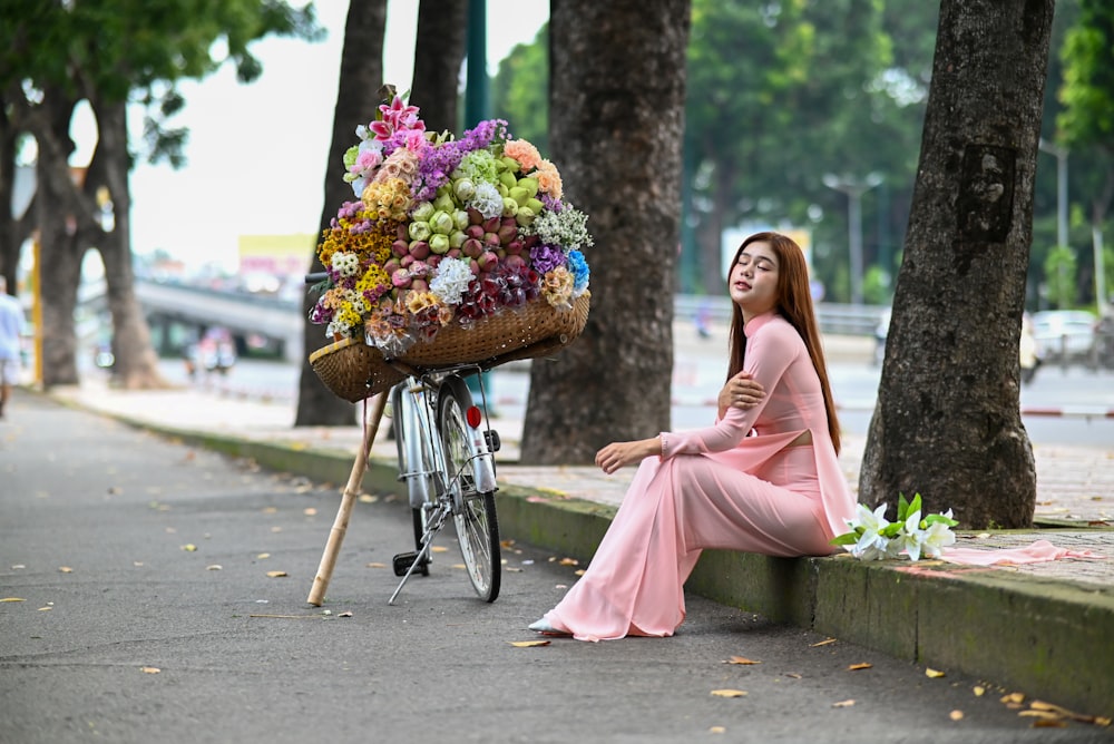 a woman sitting on a curb next to a bike with a basket of flowers on