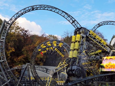 An amusement park roller coaster with intricate loops and yellow structural components. Several people are riding the roller coaster, which features tall, looping tracks and industrial design elements.