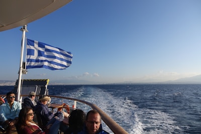 A group of people are seated on a boat under a clear blue sky, with a Greek flag waving prominently. The water is deep blue and vast, extending to a distant horizon with faint outlines of land visible.