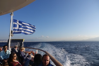 A group of people are seated on a boat under a clear blue sky, with a Greek flag waving prominently. The water is deep blue and vast, extending to a distant horizon with faint outlines of land visible.