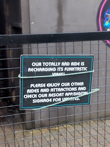 A metal fence supports a sign with a playful message about a ride being unavailable due to recharging its 'funktastic vibes'. The sign invites guests to enjoy other rides and attractions and to check the resort app or digital signage for updates.