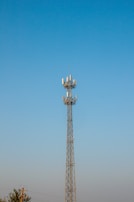 A tall communication tower with multiple antennas is set against a clear blue sky. The structure is metallic and intricately designed to support various equipment. There are some trees and power lines visible at the bottom of the image, suggesting the tower is in an open area.