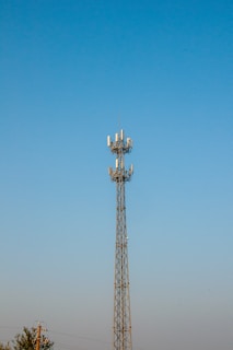 A tall communication tower with multiple antennas is set against a clear blue sky. The structure is metallic and intricately designed to support various equipment. There are some trees and power lines visible at the bottom of the image, suggesting the tower is in an open area.
