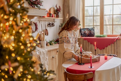 A cozy, festively decorated room featuring a woman in a traditional embroidered dress. She is standing beside a table with a red tablecloth, preparing bread while surrounded by warm candlelight. In the background, a Christmas tree with glowing lights and a decorated windowsill add to the holiday ambiance.