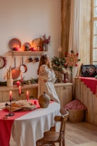 A cozy room with rustic decor featuring wooden furniture and kitchenware. A woman in a traditional embroidered dress stands by the window, surrounded by festive decorations such as candles, a small Christmas tree, and a star ornament. The table is set with a white tablecloth and a red runner, displaying a loaf of bread and a ceramic vase.