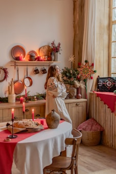 A cozy room with rustic decor featuring wooden furniture and kitchenware. A woman in a traditional embroidered dress stands by the window, surrounded by festive decorations such as candles, a small Christmas tree, and a star ornament. The table is set with a white tablecloth and a red runner, displaying a loaf of bread and a ceramic vase.