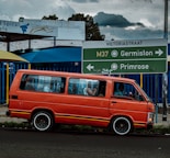 A bright orange van is parked on the side of a road with directional signs above, guiding towards Germiston and Primrose on Victoria Street. The van is set against a partly cloudy sky and some visible buildings, showcasing urban elements. A few passengers are inside the van.