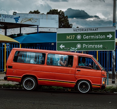 A bright orange van is parked on the side of a road with directional signs above, guiding towards Germiston and Primrose on Victoria Street. The van is set against a partly cloudy sky and some visible buildings, showcasing urban elements. A few passengers are inside the van.