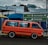 A bright orange van is parked on the side of a road with directional signs above, guiding towards Germiston and Primrose on Victoria Street. The van is set against a partly cloudy sky and some visible buildings, showcasing urban elements. A few passengers are inside the van.