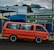 A bright orange van is parked on the side of a road with directional signs above, guiding towards Germiston and Primrose on Victoria Street. The van is set against a partly cloudy sky and some visible buildings, showcasing urban elements. A few passengers are inside the van.