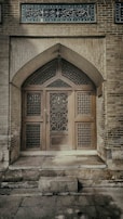 An ornate wooden door set within an arched stone frame, featuring intricate geometric patterns. Above the door, there is a set of decorative tiles with Arabic script. The stonework around the door is crafted from aged bricks, giving the structure a historic and cultural appearance.
