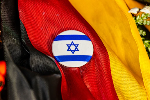 A circular button featuring the Star of David with blue and white stripes is positioned against a backdrop of vibrant fabric in the colors of black, red, and yellow.