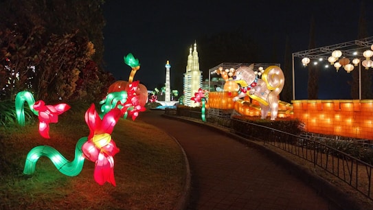 A nighttime scene featuring brightly lit, colorful lantern displays shaped like flowers and mythical creatures. The path curves around the illuminated installations, which include large animals and vibrant floral designs. In the background, architectural structures and more lanterns enhance the festive atmosphere.