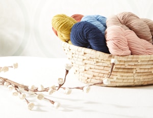 A wicker basket is filled with several skeins of colorful yarn, displayed against a soft, pastel background. The yarn colors include yellow, blue, navy, pink, and beige. In the foreground, a decorative branch adorned with small, white, rose-like flowers extends across the surface.