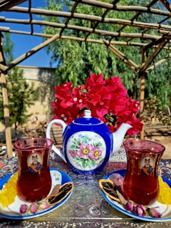 A decorative blue and white teapot with floral designs is centered on a metallic tray that has intricate patterns. Flanked on either side are two glasses filled with tea, each placed on a blue saucer and accompanied by various snacks, including sugar sticks, dried fruits, and small pink buds. In the background, there is a bouquet of vibrant red flowers, and the setting appears to be outdoors with greenery and wooden pergola overhead.