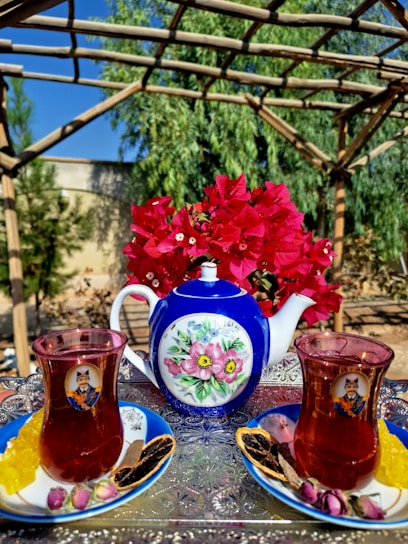 A decorative blue and white teapot with floral designs is centered on a metallic tray that has intricate patterns. Flanked on either side are two glasses filled with tea, each placed on a blue saucer and accompanied by various snacks, including sugar sticks, dried fruits, and small pink buds. In the background, there is a bouquet of vibrant red flowers, and the setting appears to be outdoors with greenery and wooden pergola overhead.