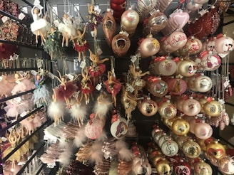 Various Christmas ornaments are displayed on shelves, including ballerina figurines in different poses and colors, and spherical baubles with intricate designs. Many of the decorations incorporate glitter and feathers, adding a festive, elegant touch.