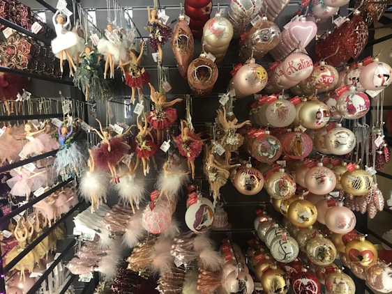 Various Christmas ornaments are displayed on shelves, including ballerina figurines in different poses and colors, and spherical baubles with intricate designs. Many of the decorations incorporate glitter and feathers, adding a festive, elegant touch.