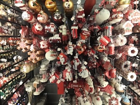 A colorful display of Christmas ornaments and decorations in various shapes and sizes, including Santa figures, snowflakes, and Coca-Cola branding. The items are arranged on shelves, creating a festive and vibrant atmosphere.