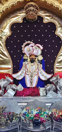 A beautifully adorned deity statue with intricate jewelry and vibrant clothing, placed at the center of a shrine surrounded by ornate decorations. Silver figurines of cows flank the statue, while a collection of colorful figurines and flowers are arranged below. The background features a dark purple velvet-like fabric with golden accents.