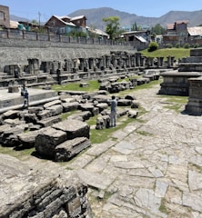Ancient stone ruins scattered across a grassy area with a background of residential houses and distant mountains. Two people are exploring the ruins, taking photographs and observing the historical site.