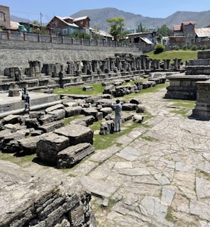 Ancient stone ruins scattered across a grassy area with a background of residential houses and distant mountains. Two people are exploring the ruins, taking photographs and observing the historical site.